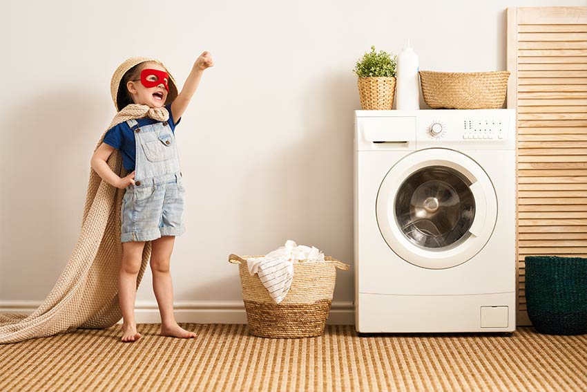 Child in superhero costume near washing machine and laundry basket, symbolizing playful cleaning.